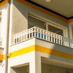 A modern residential building with a sunlit balcony, featuring geometric railings and bold color accents.